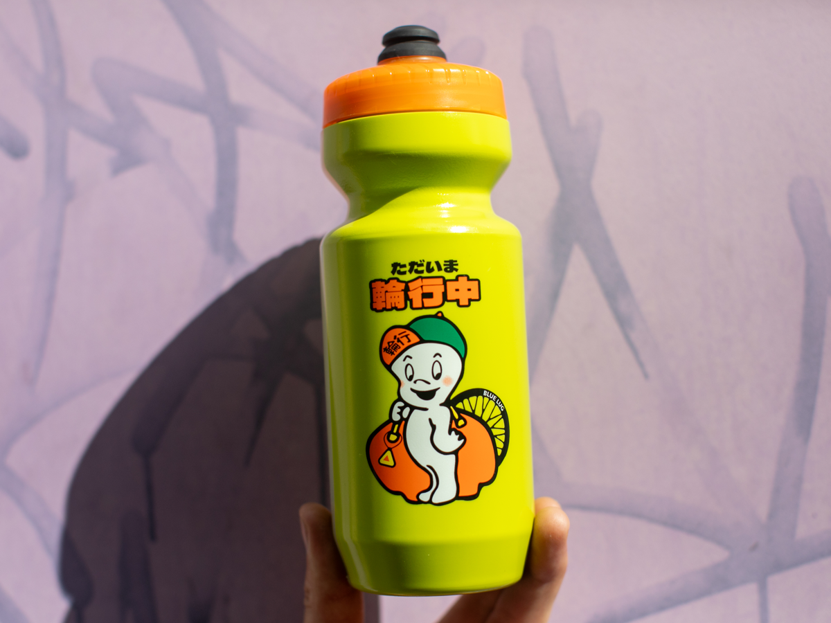 Blue Lug Rinko-kun Bottle in Yellow Green with orange cap — Rinko-kun mascot carrying a big rinko bag with a wheel sticking out, Japanese text reading "currently doing rinko"