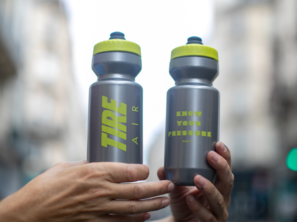 Blue Lug Tire Air water bottle, both sides shown. Left side reads "TIRE AIR" in bold neon yellow lettering; right side reads "KNOW YOUR PRESSURE" with the Blue Lug logo below. Grey body with neon yellow cap.