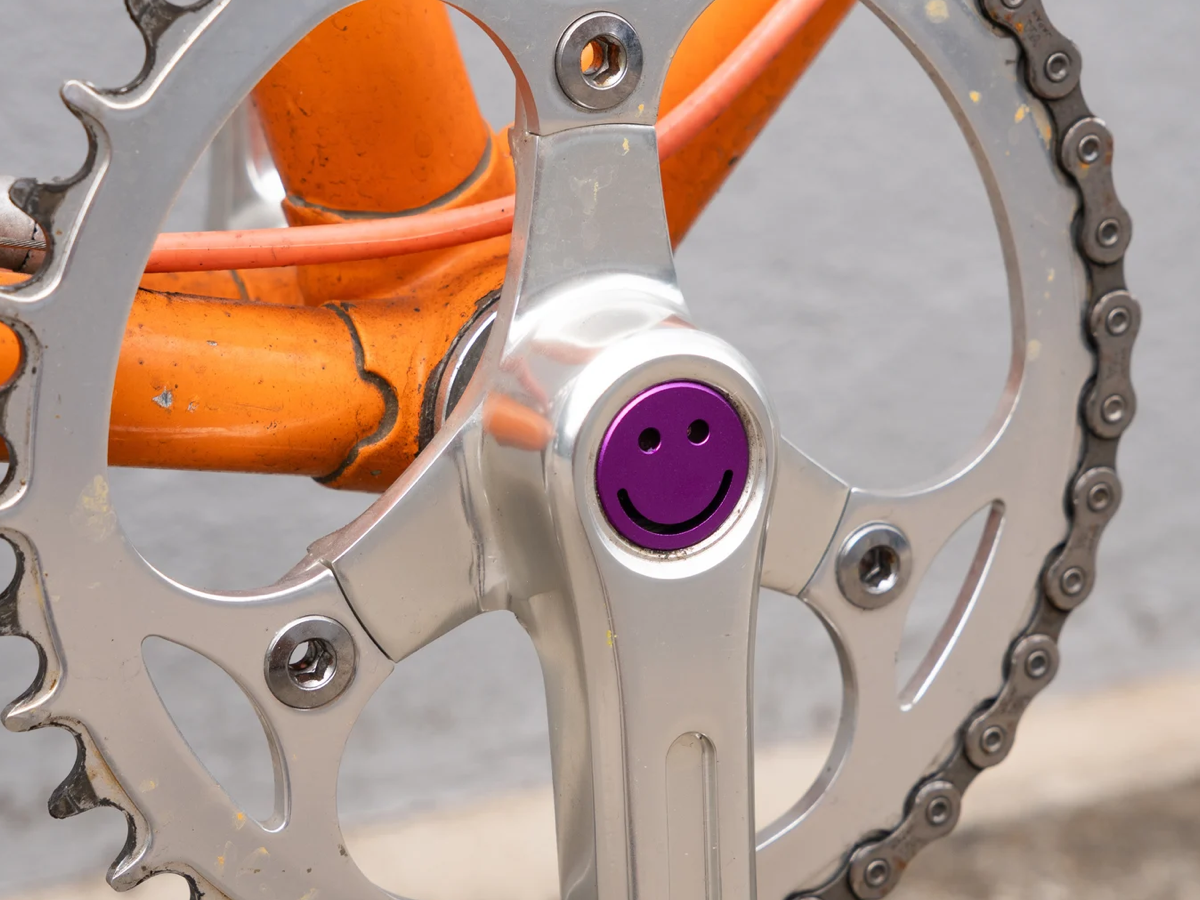 Purple Bike Felix Smiley aluminium crankset cap fitted to a silver double chainring crankset on an orange bike