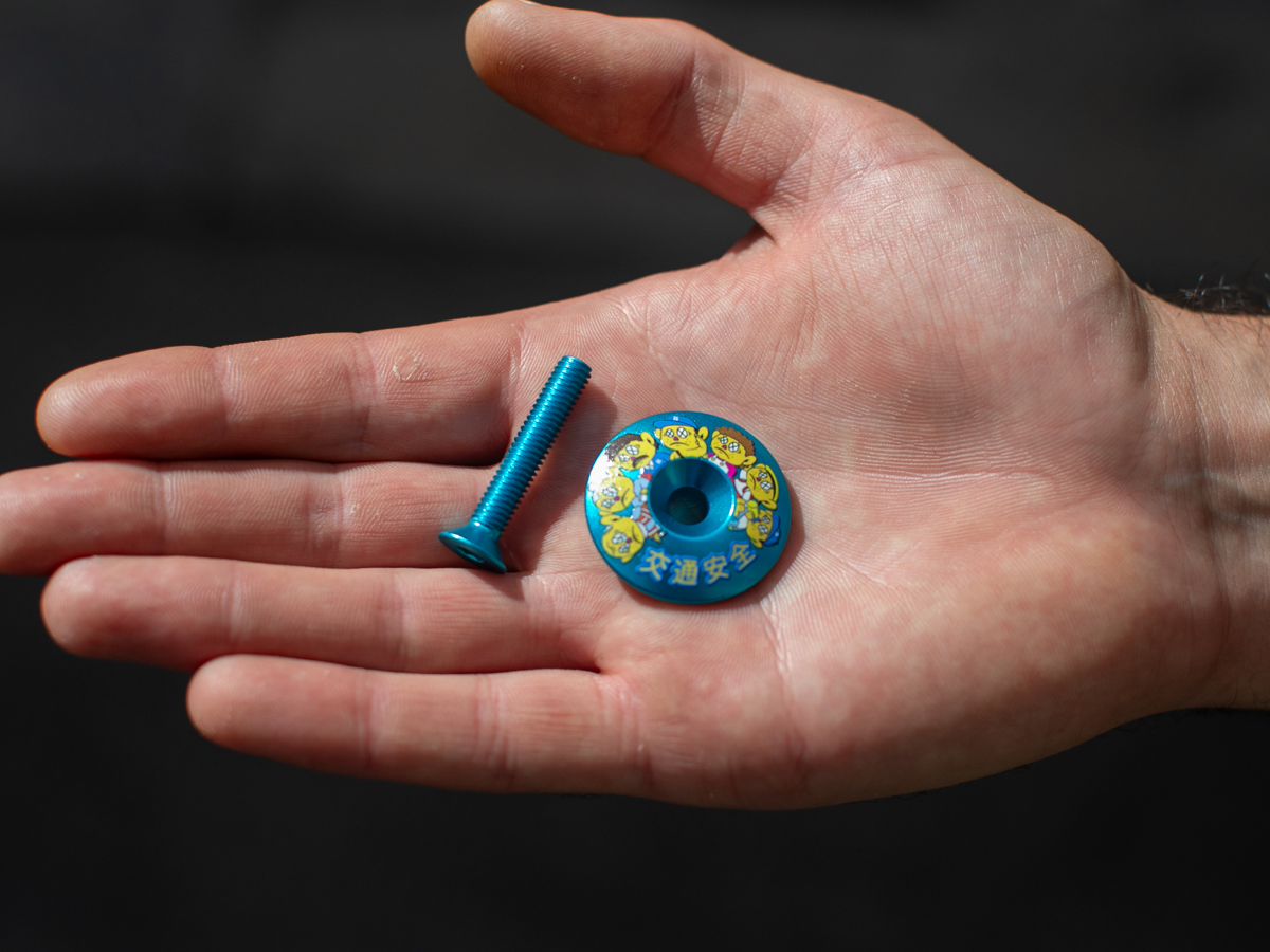 Blue Lug Billboard Stem Cap, Anzen Family variant — teal anodized aluminum cap with Blue Lug mascot characters and "交通安全" (road safety) text, with matching teal bolt, held in a palm