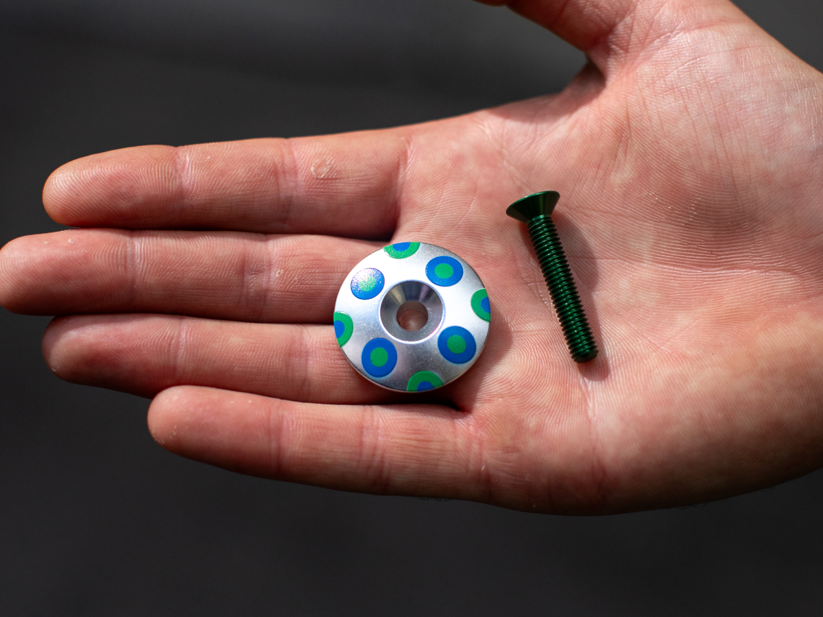 Blue Lug Billboard Stem Cap, Dots variant — silver aluminum cap with blue and green polka dot pattern, with dark green bolt, held in a palm