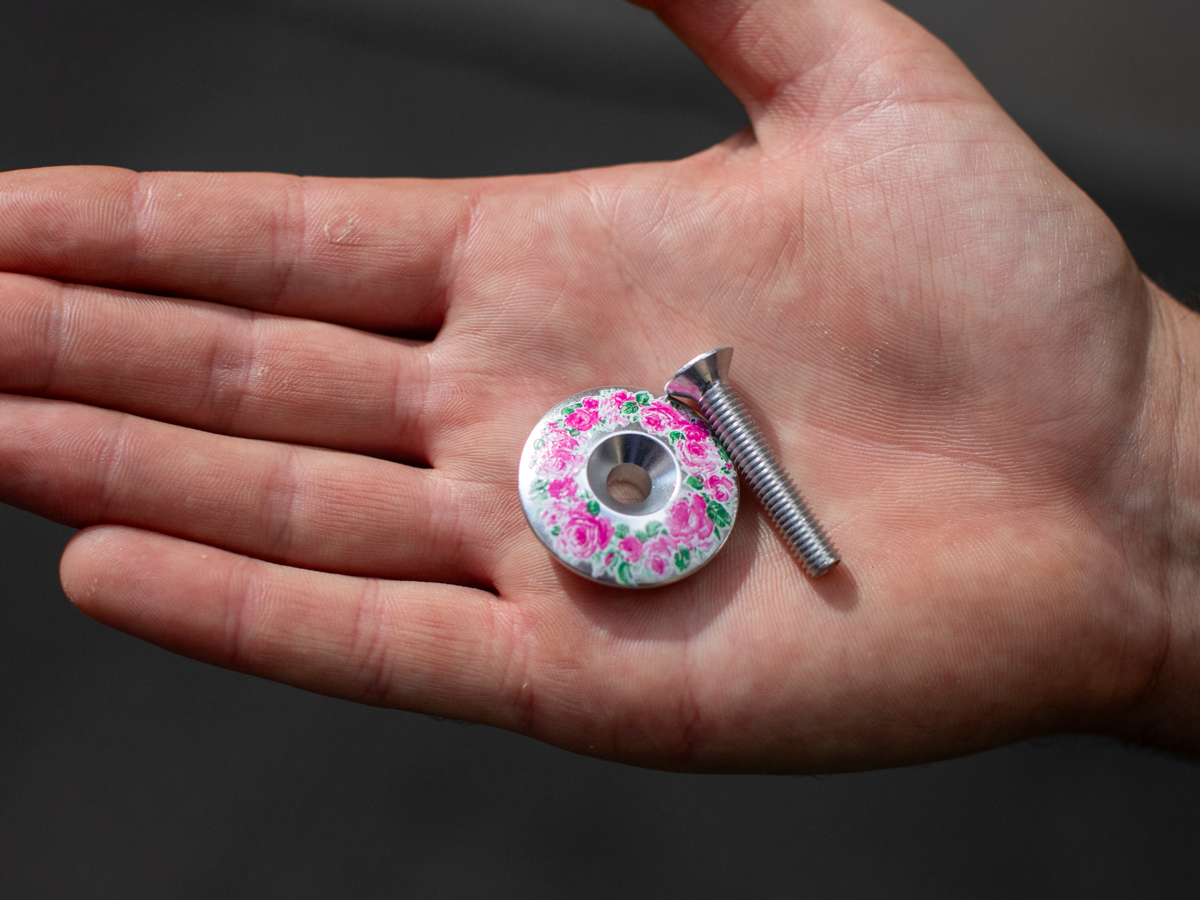 Blue Lug Billboard Stem Cap, Floral variant — silver aluminum cap with pink rose floral pattern, with silver bolt, held in a palm