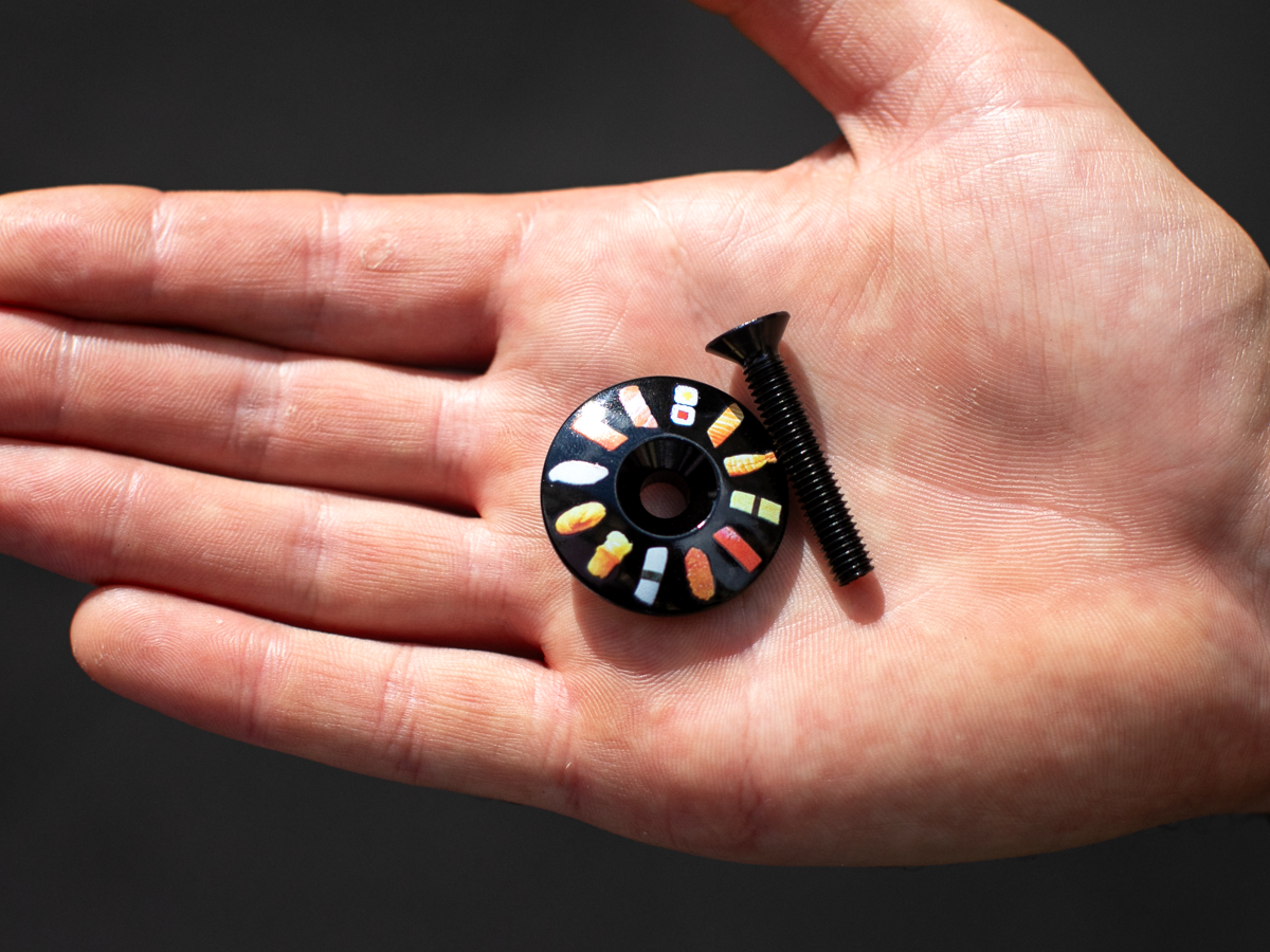 Blue Lug Billboard Stem Cap, Sushi variant — black aluminum cap with colorful sushi illustrations arranged in a circle, with black bolt, held in a palm