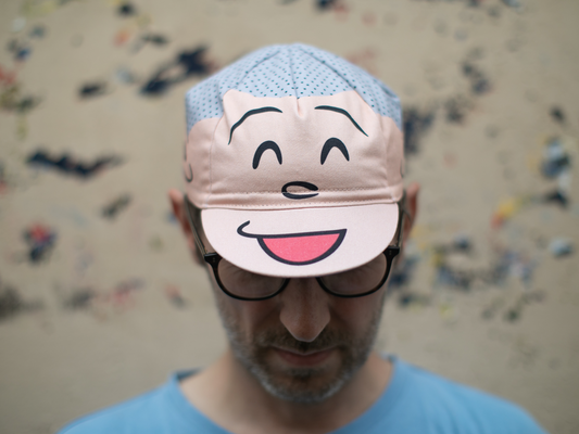 Casquette de cyclisme Blue Lug Good Boy portée visière baissée, vue de face. Les panneaux avant affichent un grand visage souriant en rose avec des yeux plissés et une bouche ouverte, sur un fond bleu clair à pois.