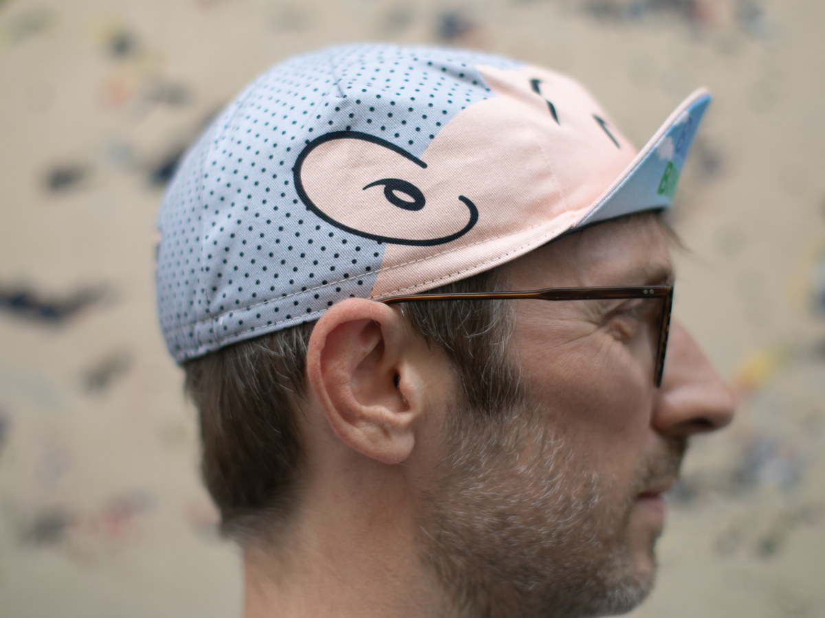 Side profile of the Blue Lug Good Boy Cycle Cap, showing the pink cartoon face panel on the front and the light blue polka dot panel on the crown, with the classic pointed cycling cap brim.
