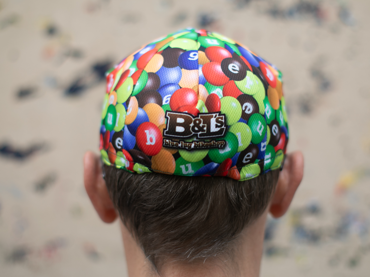 Rear view of the Blue Lug Cycling Cap in Chocolate/Poly, showing the "B&L's Blue Lug Bikeshop" logo patch styled as a parody of the M&M's brand.