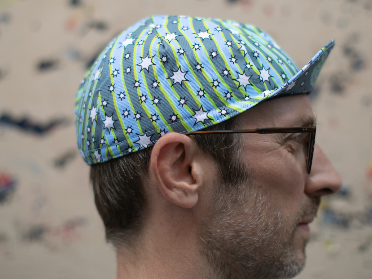 Profil de la casquette de cyclisme Blue Lug Dreamer, montrant le motif de bandes tourbillonnantes et d'étoiles en bleu, gris et jaune-vert fluo, avec la visière pointue caractéristique.