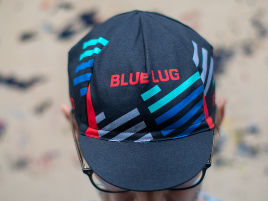 Casquette de cyclisme Blue Lug Original coloris noir, vue de dessus. Le motif de bandes géométriques en cyan, bleu, gris et rouge avec le texte "BLUE LUG" en rouge est visible sur les panneaux.