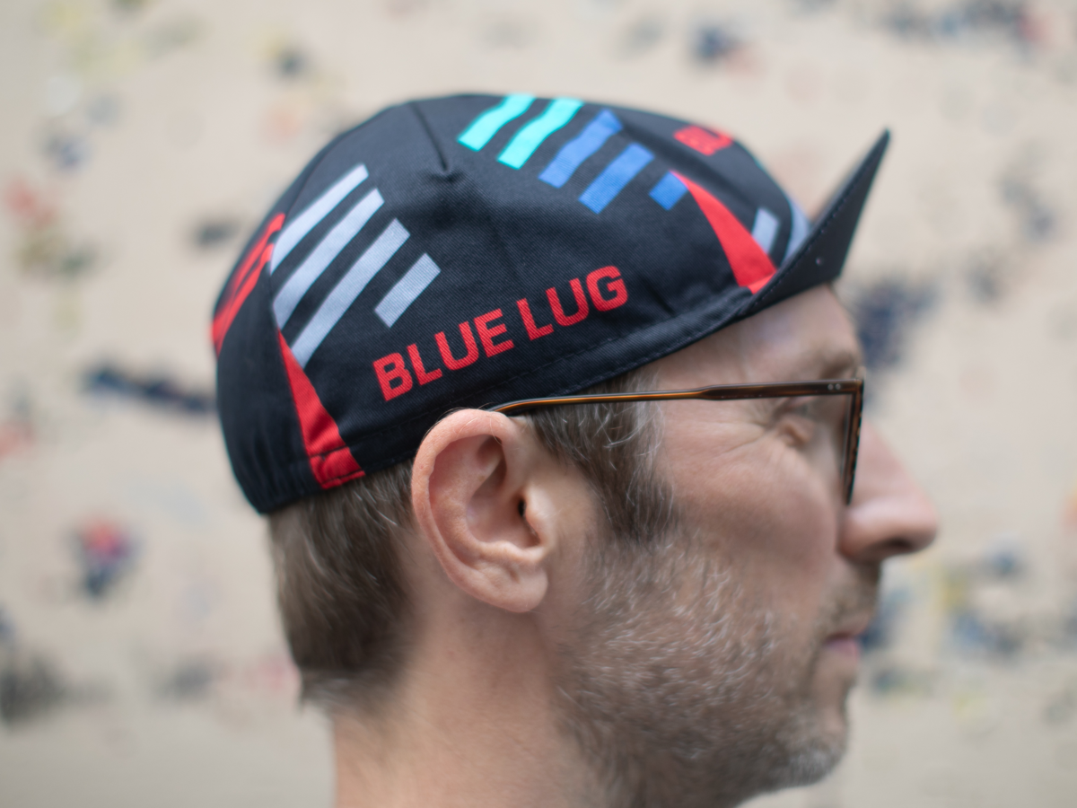 Side profile of the Blue Lug Original Cycle Cap in black, showing the geometric stripe design and "BLUE LUG" text in red, with the classic pointed cycling cap brim.