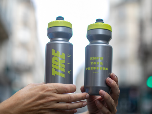 Bidon Tire Air de chez Blue Lug, les deux faces visibles. La face gauche affiche "TIRE AIR" en grandes lettres jaune fluo ; la face droite affiche "KNOW YOUR PRESSURE" avec le logo Blue Lug en dessous. Corps gris avec bouchon jaune fluo.