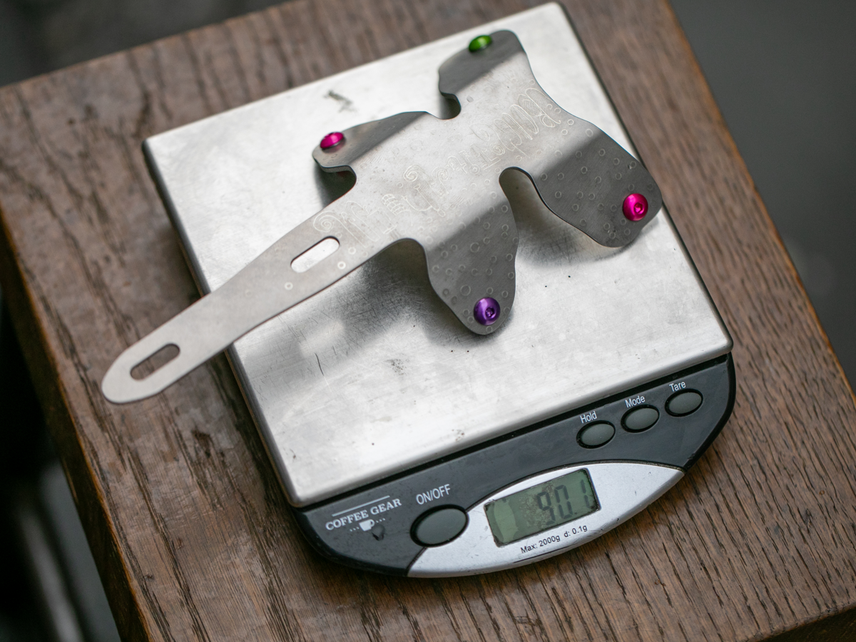 Botched Bikes Titanium Dragonfly sur une balance affichant 90,1g, montrant le profil coudé à 45° et les oreilles de fixation de porte-bidon. Boulons anodisés rose et violet.