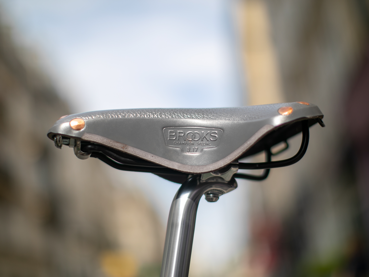Brooks x Rivendell B17 saddle in gray — side view on a silver seatpost, showing the pebble leather top, copper rivets at the nose, carved lower flap and "Brooks Champion Special B17" embossed stamp