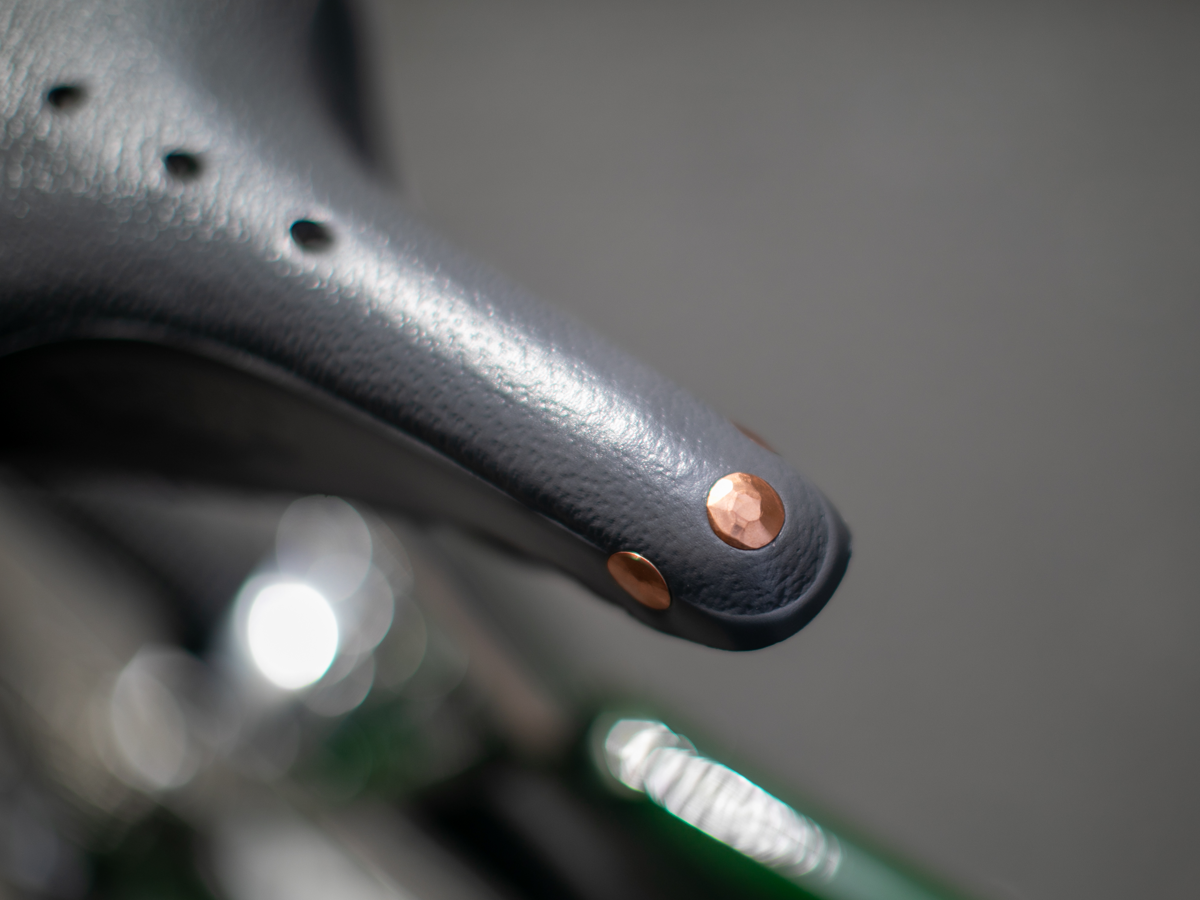 Selle Brooks x Rivendell B17 — gros plan sur les deux rivets cuivre du flanc arrière sculpté, avec cadre de vélo vert visible en dessous