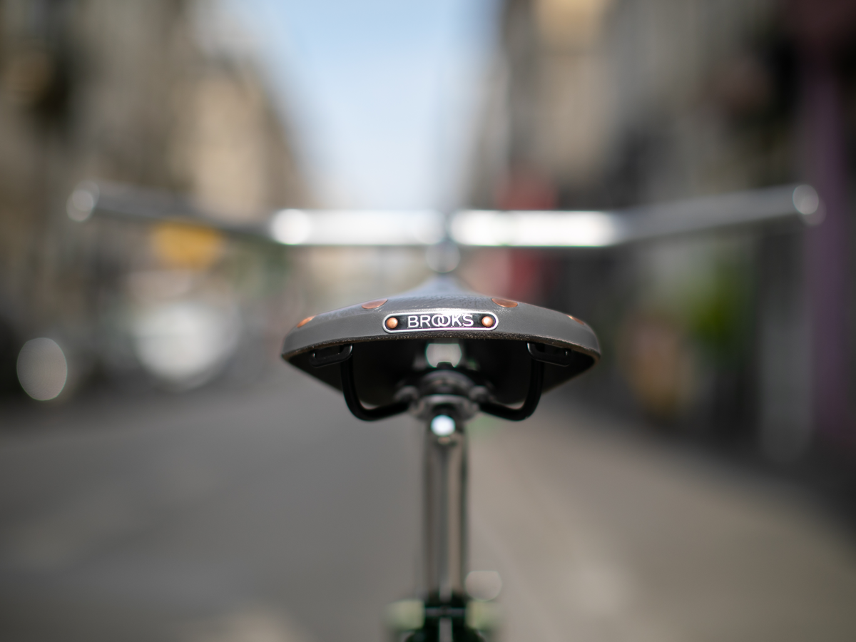 Selle Brooks x Rivendell B17 — vue de derrière montrant le badge Brooks avec rivets cuivre, rails acier noir et tige de selle, rue parisienne en arrière-plan