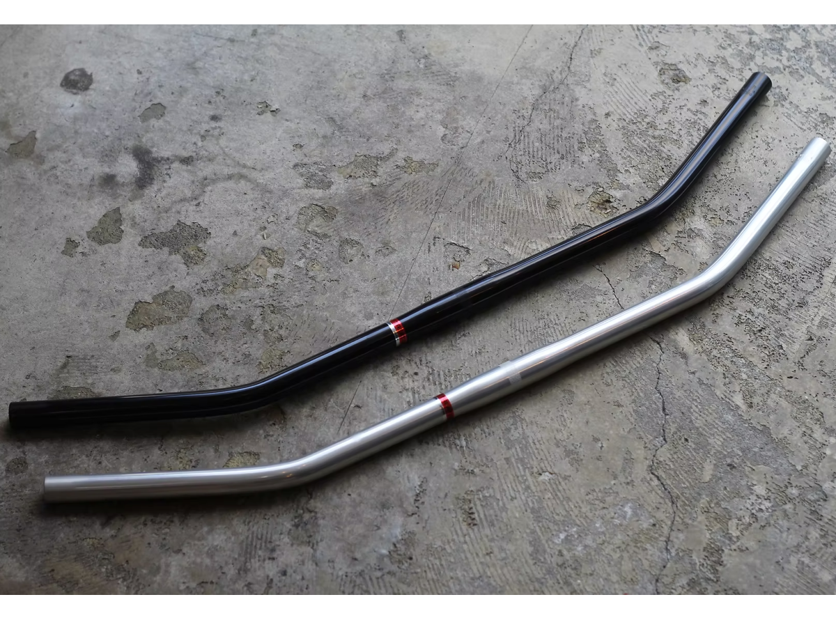 Magic Components x Nitto Carter Bar in black and raw aluminum, laid side by side on a concrete floor. The gentle 25° backsweep and red center accent ring are visible on both colorways.