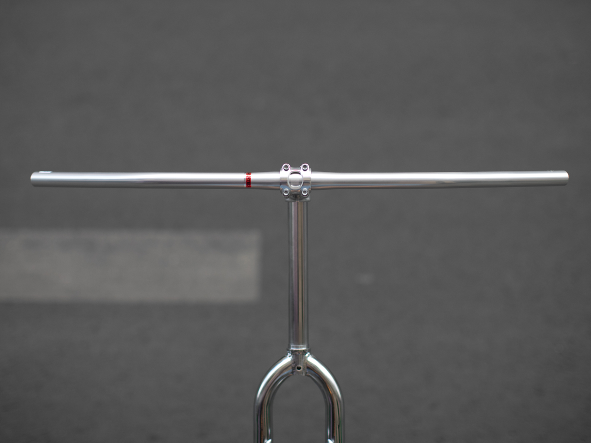 Carter Bar in raw aluminum, front view, mounted on a display stand with a threadless stem. The full 750mm width and subtle backsweep are visible against a grey background.