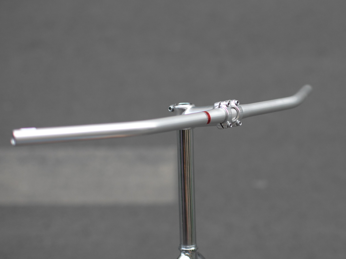 Carter Bar in raw aluminum, three-quarter view showing the 25° backsweep. The red center accent ring and stem clamp detail are visible in the foreground.