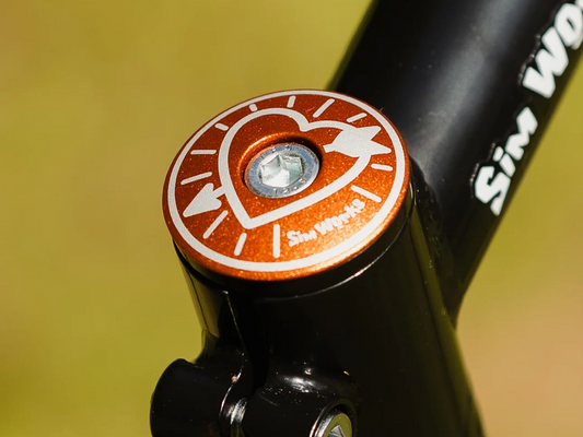 SimWorks Heart & Arrow Stem Cap in Sunrise orange — installed on a black stem, showing the heart and arrow motif with SimWorks logo engraved in white