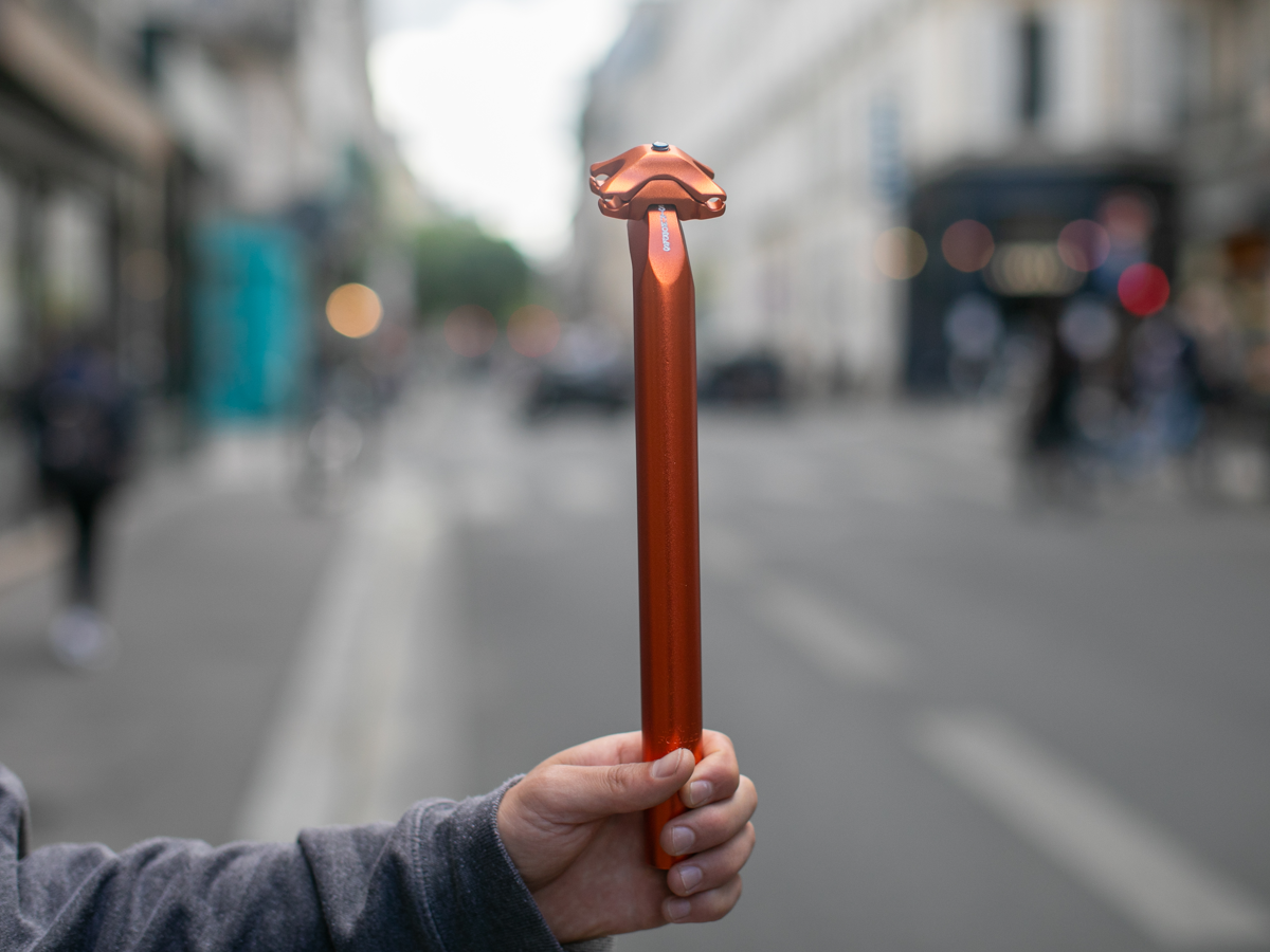 Tige de selle SimWorks Beatnik Sunrise coloris Sunrise orange — vue complète de la tige en aluminium forgé 300mm avec tête de serrage, tenue devant une rue parisienne floue