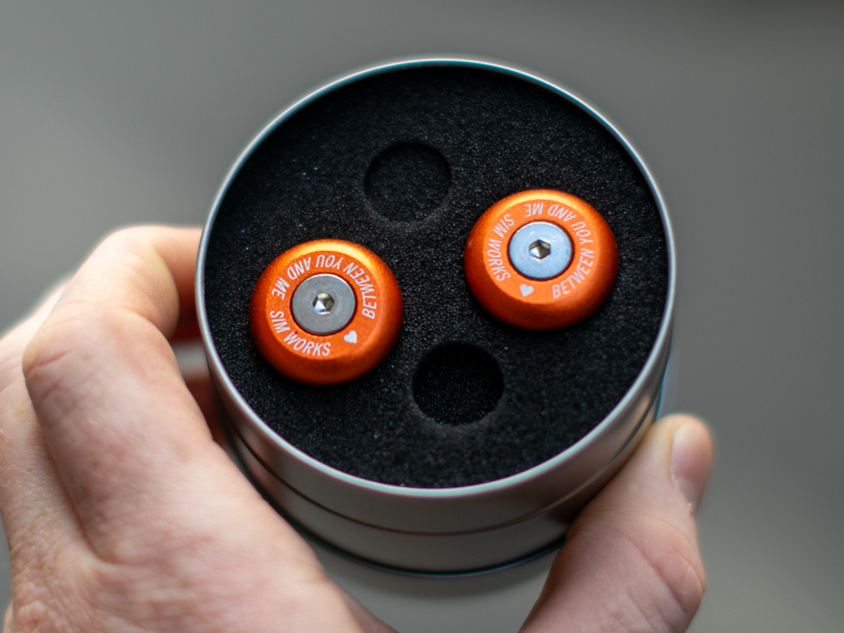 SimWorks Sim Drops Bar Ends in Sunrise orange — pair shown in round metal tin with black foam insert, engraved "Between You and Me / Sim Works" text visible on the face