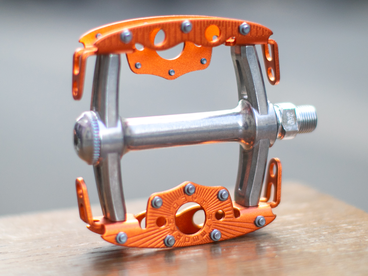 Pédale SimWorks Taco coloris Sunrise orange — profil de côté montrant la structure de cage ouverte, les picots d'adhérence, l'axe argenté avec réglage de roulement à cônes et cuvettes, et la gravure SimWorks en soleil sur la plateforme