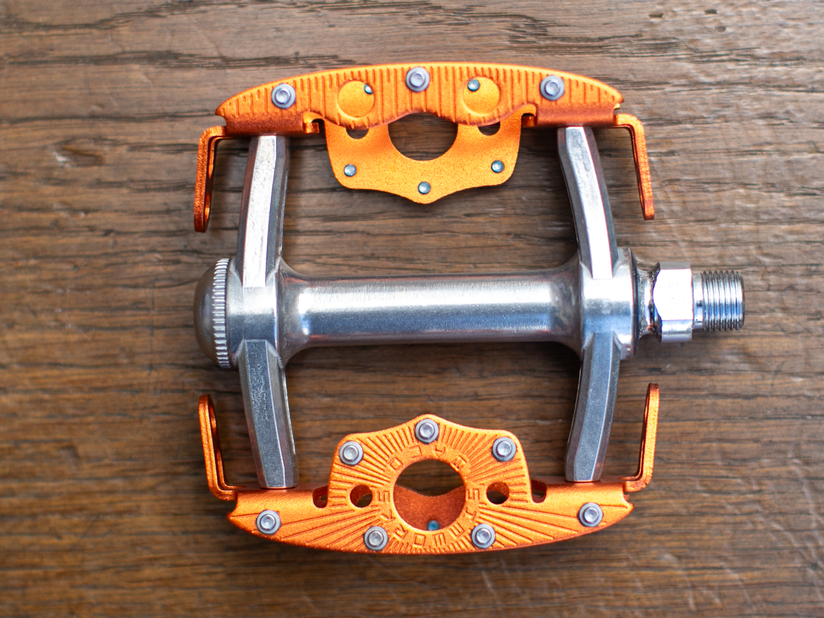 Pédale SimWorks Taco coloris Sunrise orange — vue de dessus sur bois montrant la plateforme complète, les picots, la cage ouverte et la gravure SimWorks en soleil au centre