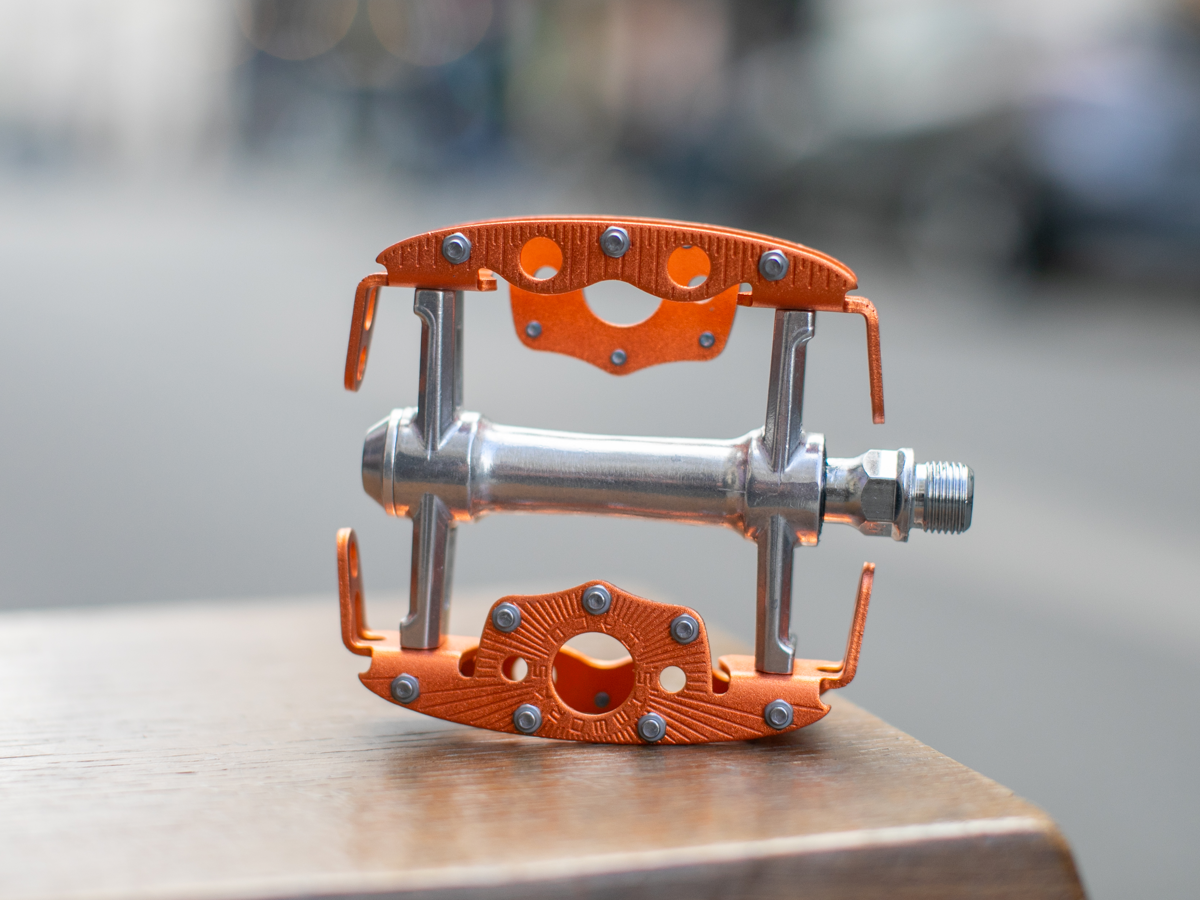 Pédale SimWorks Taquito coloris Sunrise orange — profil de côté sur bois montrant la structure de cage compacte et ouverte, les picots, l'axe CrMo et la gravure SimWorks en soleil sur la plateforme