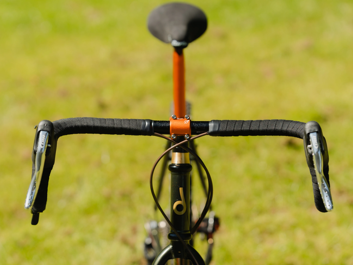 Plaque frontale SimWorks Tomboy coloris Sunrise orange — montée sur une potence noire avec cintre courbé et guidoline sombre, sur un vélo devant un fond vert, avec la tige de selle Beatnik Sunrise visible derrière