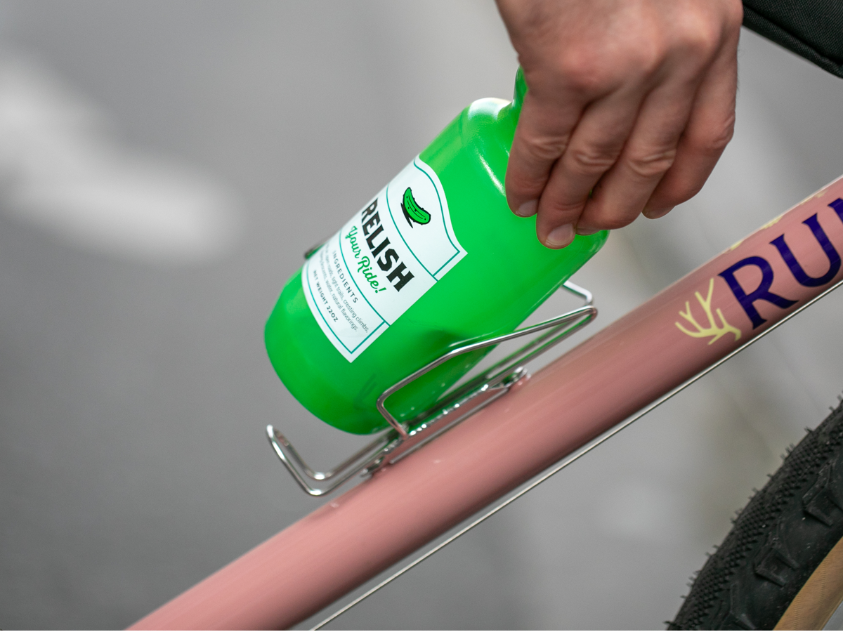 Hand pulling a water bottle out of a Velo Orange Crescent sideloading bottle cage on a pink bike frame